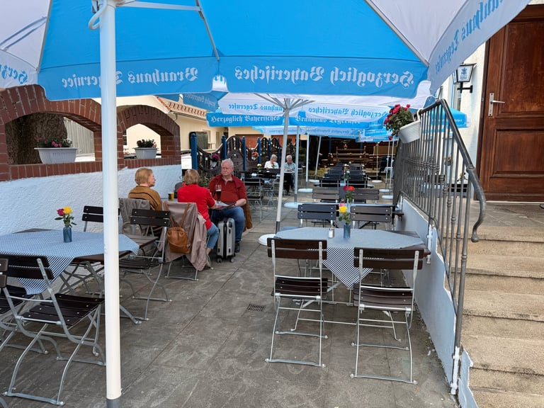 Outdoor restaurant patio with blue umbrella, white tables and chairs, customers seated, stone courtyard