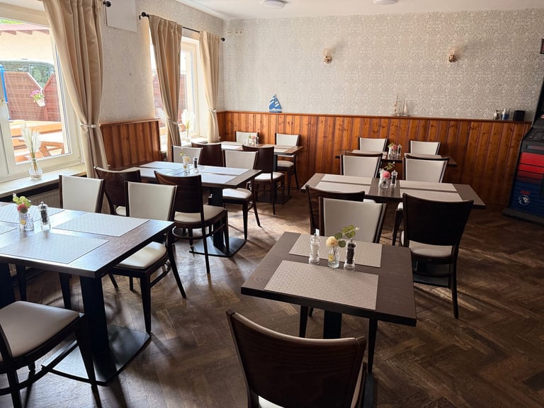 Empty restaurant dining room with wooden tables, black chairs, wood paneling, and large windows with curtains